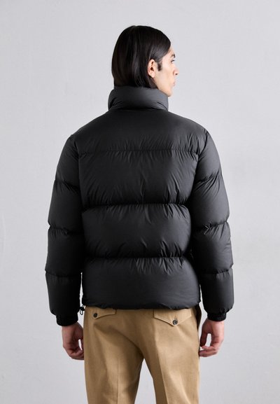 Doudoune noire avec un col montant, design matelassé et poignets élastiques. Portée sur un pantalon beige, mettant en avant une texture mate.