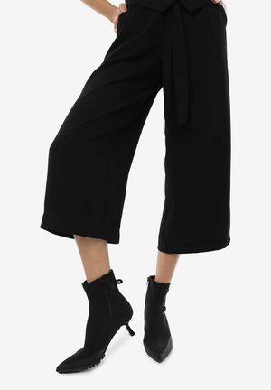 Pantalon large cropped noir avec une ceinture à nouer et des bottes noires arrivant à la cheville, dotées d'un bout pointu et d'une fermeture éclair. Texture de tissu lisse.