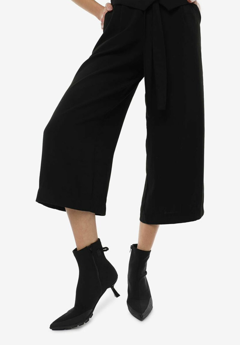 Zwarte cropped broek met wijde benen, een strikceintuur en enkelhoge zwarte laarzen met een spitse neus en ritssluiting. Glad textuur van de stof.