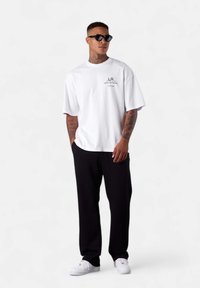 White oversized t-shirt with black text, paired with black pants and white sneakers. Model also wears black sunglasses and has tattoos.