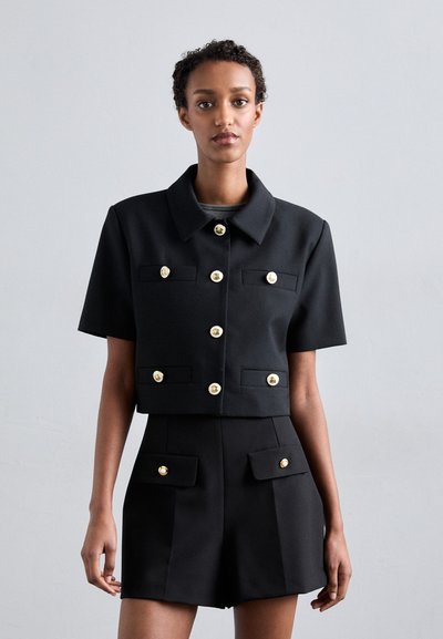 Sort cropped jakke med korte ærmer og guldknap detaljer, parret med high-waisted shorts med lignende knapper og lomme detaljer.