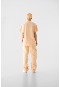 Világos narancssárga, rövid ujjú póló hálós textúrával. Világos narancssárga cargo nadrággal, oldalzsebekkel. Fehér tornacipő.