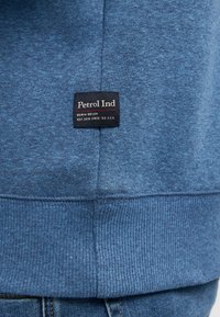 Blauwe sweatshirt van een textuurstof. Heeft een zwart label met de tekst "Petrol Ind" en rode accenten. Ribgebreide manchetten en hem ontwerp.