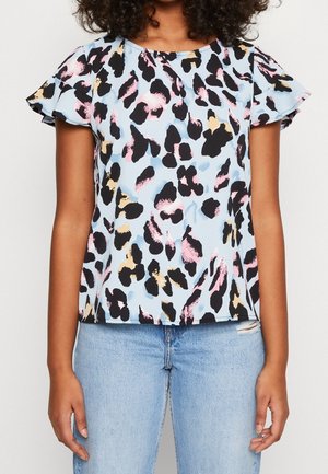 Woman wearing a short-sleeve, light blue blouse with a black, pink, and beige abstract pattern and light blue ripped jeans.