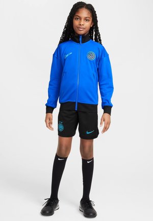 Tenue de sport Nike comprenant une veste zippée bleue avec des accents noirs et un logo, un short noir avec des détails bleus, des chaussettes noires et des chaussures noires.