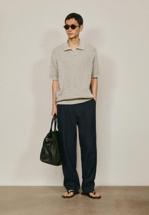 Homme portant un polo en maille beige à manches courtes, pantalon ample foncé, lunettes de soleil, tongs, tenant un sac cabas en cuir noir, debout contre un mur uni.