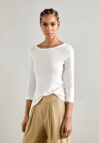 Top blanco de manga larga con escote barco, hecho de una tela suave, combinado con pantalones de talle alto beige con pliegues. Diseño simple, sin patrones.