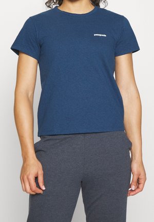 Persoon die een nauwsluitend donkerblauw Patagonia T-shirt en grijze broek draagt, staand met ontspannen armen langs het lichaam tegen een effen achtergrond.