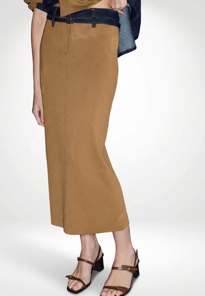 Žena v hnědé midi sukni a hnědých sandálech na blokovém podpatku se střihem přes nohy, drží u pasu džínovou bundu.