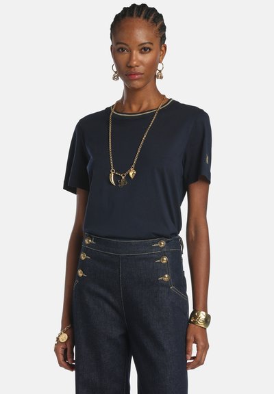 Fonott hajú nő, aki tengerészkék pólót, magas derekú sötét farmernadrágot arany gombokkal, valamint arany ékszereket, beleértve nyakláncot, fülbevalókat és karkötőt visel.