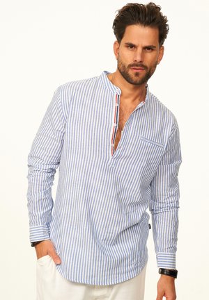 Mann trägt ein blau-weiß vertikal gestreiftes Hemd mit Stehkragen und Knöpfen, weiße Hose, Armbanduhr und Armband, posiert mit der Hand in der Tasche.