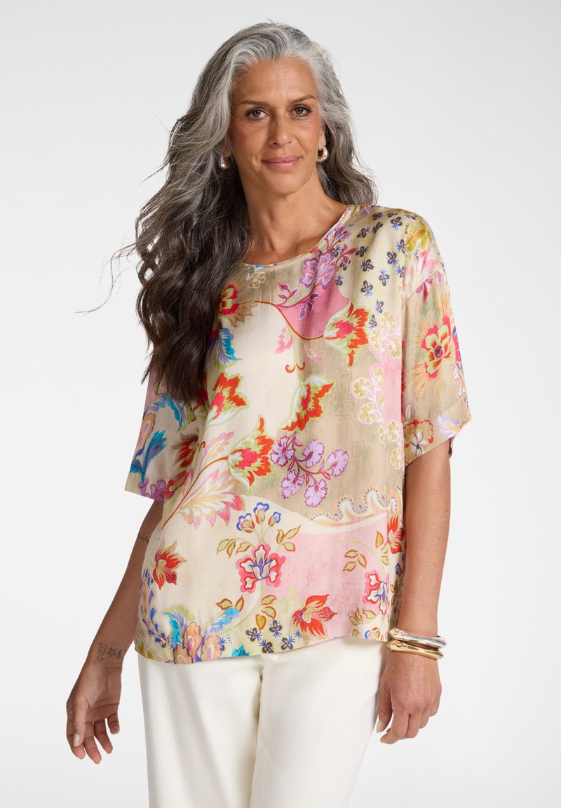 Mujer con cabello largo y gris que lleva una blusa floral colorida y pantalones blancos, complementada con brazaletes dorados y pendientes de aro.
