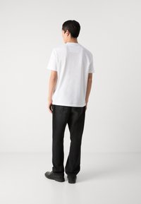 Weißes kurzärmliges T-Shirt aus Baumwolle, mit Rundhalsausschnitt und einem kleinen Etikett auf der Rückseite. Tragen mit schwarzen geraden Hosen und schwarzen Schuhen.