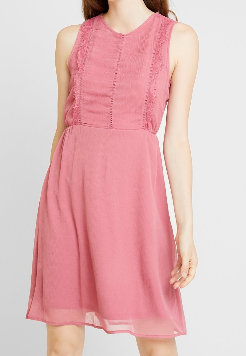 Robe rose sans manches longueur genou avec bordure en dentelle sur le corsage portée par une femme debout devant un fond clair uni.