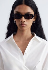 Femme aux longs cheveux foncés portant des lunettes de soleil ovales noires, des boucles d’oreilles anneaux argentées et une chemise blanche boutonnée sur un fond uni.