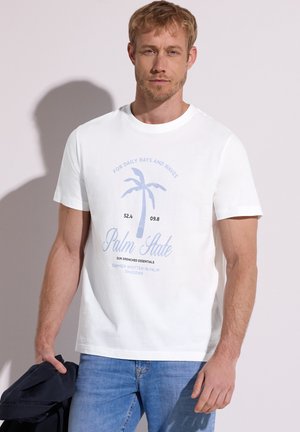 Homme blond portant un t-shirt blanc avec un motif de palmier bleu et du texte, tenant une veste sombre dans sa main gauche sur un fond uni.