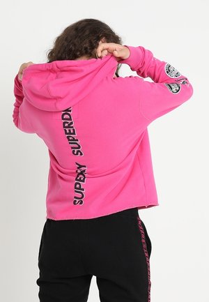 Persoon die de capuchon van een felroze hoodie met de tekst "SUPERDRY" op de achterkant omhoog trekt, draagt een zwarte broek met roze zijstrepen.