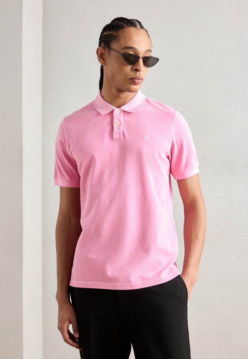Rosa polo skjorte med klassisk krage, tre-knappslukking og en liten logo. Brukt med svarte bukser og solbriller. Glatt bomullstekstil.