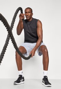 Haut de sport sans manches noir, short gris clair, baskets noir et blanc, tenant une corde de bataille noire texturée, sur un fond blanc.