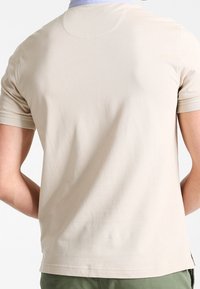 Male figure wearing a beige short-sleeve shirt, viewed from the back. Soft fabric appearance and relaxed fit, standing against a neutral background.