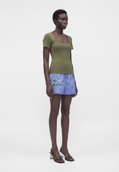 Γυναίκα που στέκεται, φοράει ελιάς πράσινη κοντομάνικη μπλούζα, μπλε και λευκές ριγέ φλοράλ σορτς και μαύρα σανδάλια με τακούνι σε απλό φόντο.