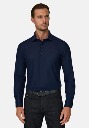 Homme portant un polo bleu marine à manches longues rentré dans un jean foncé avec une ceinture noire, debout devant un fond uni.