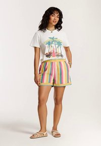 Hvid t-shirt med korte ærmer og et farverigt grafisk design, kombineret med flerfarvede stribede shorts i gul, pink og blå. Afslappede sandaler.