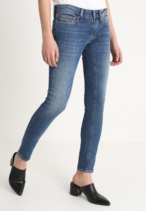 Jean bleu slim porté avec des mules à talons noires et un haut blanc, montrant les jambes et les mains avec des bagues sur un fond uni.