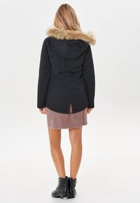 Giacca nera con cappuccio e bordatura in pelliccia sintetica, con vita regolabile, apertura sul retro e un vestito lavorato a maglia rosa sotto. Stivali neri alla caviglia.