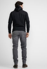 Sudadera negra con cordón, combinada con jeans gris claro. Modelo visto desde atrás, de pie sobre un fondo sólido claro.