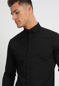 Schwarzes Langarmhemd mit Knopfleiste, schmalem Kragen, subtilen Glanz und figurbetontem Design, mit schwarzen Knöpfen auf der Vorderseite.