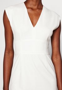 Vitt klänning med djup V-ringning, korta ärmar och figurnära midja. Tyget har en slät yta med subtila sömdetaljer.