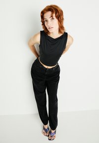 Černý bezrukávový top s volným výstřihem, spárovaný s volnými černými kalhotami. Modelka má na sobě purpurové žabky. Jednoduchý, nezdobený design.