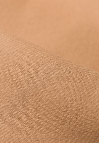 Tela color beige con un patrón de espiga, exhibiendo una textura suave y un ligero brillo, enrollada en el borde para dar dimensión.