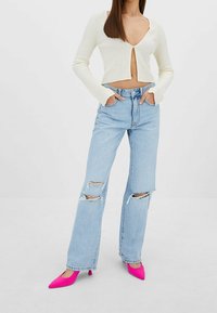 Bílý vypasovaný zkrácený kardigan s hlubokým výstřihem do V a knoflíkovým zapínáním, v kombinaci s světlomodrými roztrhanými širokými džíny a jasně růžovými podpatky.