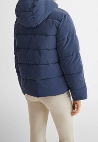 Osoba v tmavě modré prošívané bundě s kapucí a béžových kalhotách, stojící zády k fotoaparátu před světlým pozadím.