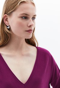 Maglione fucsia a V in morbido tessuto lavorato a maglia, con una vestibilità comoda e dettagli a coste sul collo. Orecchini a cerchio argentati completano il look.