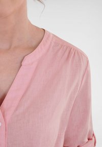 Light pink blouse with a round neckline, button closure, and gathered sleeve detail. Made of lightweight fabric with a slightly textured finish.