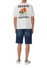Bijela pamučna majica s tiskanim grafičkim prikazom papaje i natpisom "PAPAYA okus SURFWAX." Uparena s trapericama i teniskama.