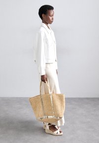 Bolso de paja con textura tejida, detalles metálicos y dos asas resistentes, llevado por una persona que viste una chaqueta blanca y una falda crema.