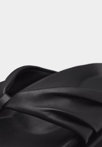 Κοντινό πλάνο μαύρου σανδαλιού με φαρδιά, απαλή, στριφογυριστή λουρίδα και διαμορφωμένη σόλα, σε απλό ανοιχτό φόντο.