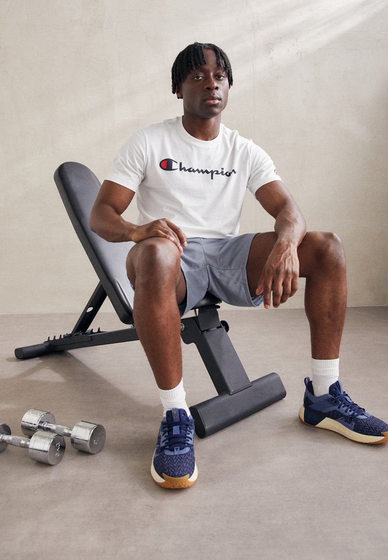 Camiseta blanca de manga corta de Champion, pantalones cortos deportivos grises, zapatillas de deporte azules con textura, sentado en un banco de entrenamiento negro ajustable, con mancuernas cerca.