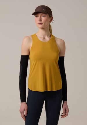 Femme portant un haut sans manches moutarde, des manchons noirs, des leggings foncés et une casquette marron sur un fond uni.