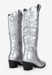 Botas de cuero sintético plateadas hasta la rodilla con una superficie texturizada, detalles de costura y un tacón cuadrado. Cuentan con una cremallera lateral para facilitar su uso.