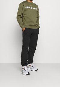 Olivově zelená mikina Nike Air s kulatým výstřihem, v kombinaci s černými tepláky a bílými sportovními botami se šedými a oranžovými detaily.