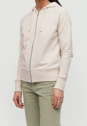 Osoba nosící světle béžovou mikinu na zip s tkaničkami a světlé olivově zelené kalhoty, stojící před jednobarevným pozadím.