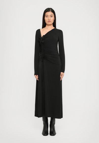 Schwarzes langärmliges Kleid mit einem gerafften Vorderteil und einem floralem Akzent auf der Schulter, mit tiefem V-Ausschnitt und ausgestellt geschnittenem Saum. Getragen mit schwarzen Stiefeln.