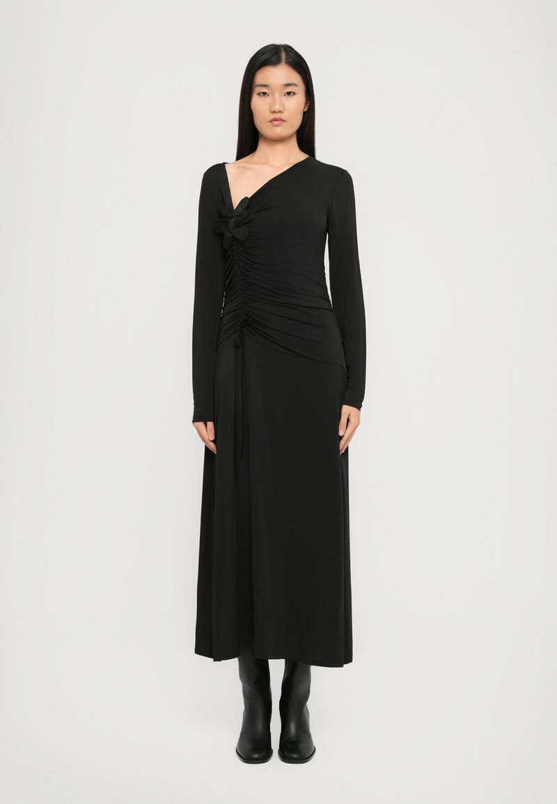 Fekete, hosszú ujjú ruha ráncolt elülső résszel és virágos akcentussal a vállon, mély V-nyakkal és bővülő szegéllyel. Fekete csizmával viselve.