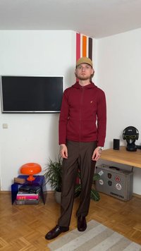 Hoodie zippé rouge en tissu texturé, logo sur la poitrine ; pantalon brun ; chaussures noires ; se tenant contre un mur blanc avec des accents colorés.