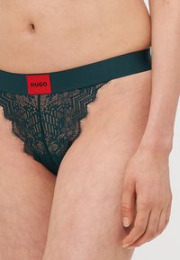 Lace thong i mørkegrønn med en myk elastisk midjebånd som har en rød logo-patch, intrikat blonde mønster og bølgete kanter.